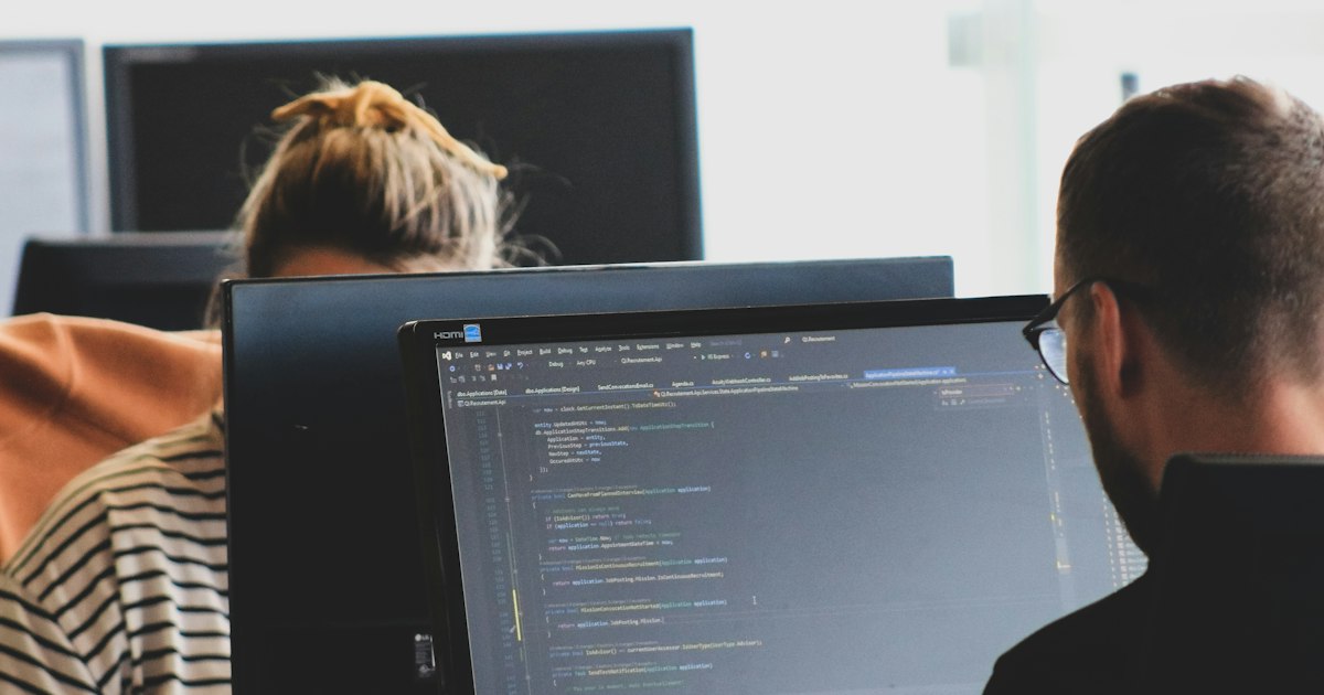Two people working on computer code at monitors in a bright office workspace