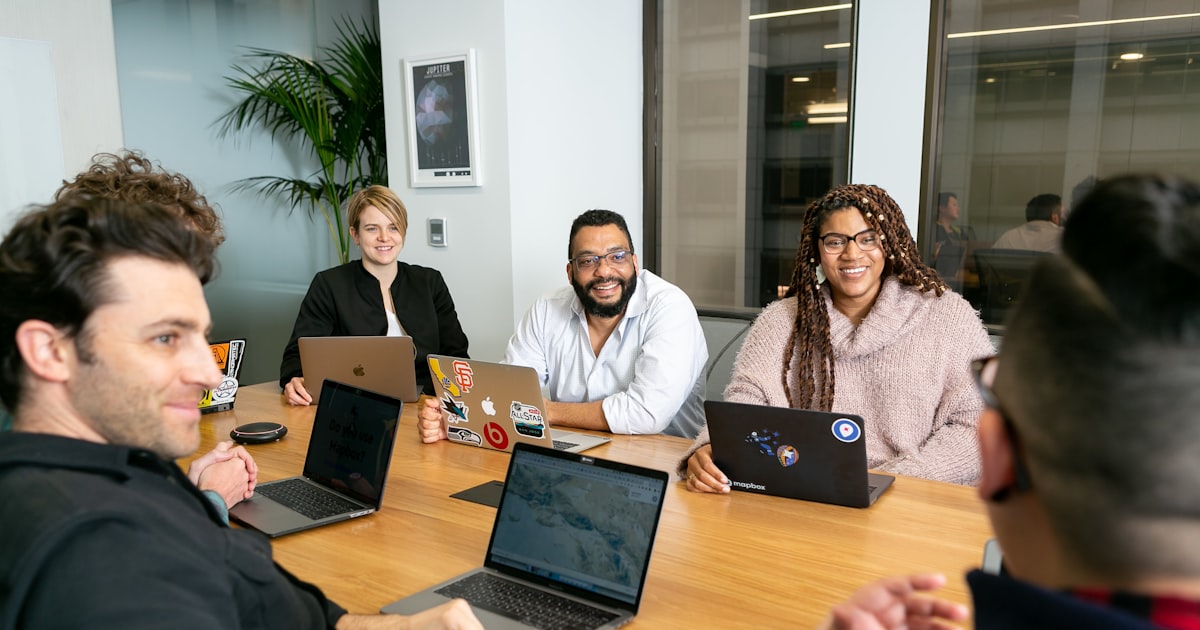 four people all on laptops, two men and two women, listen to person talking in a board meeting 