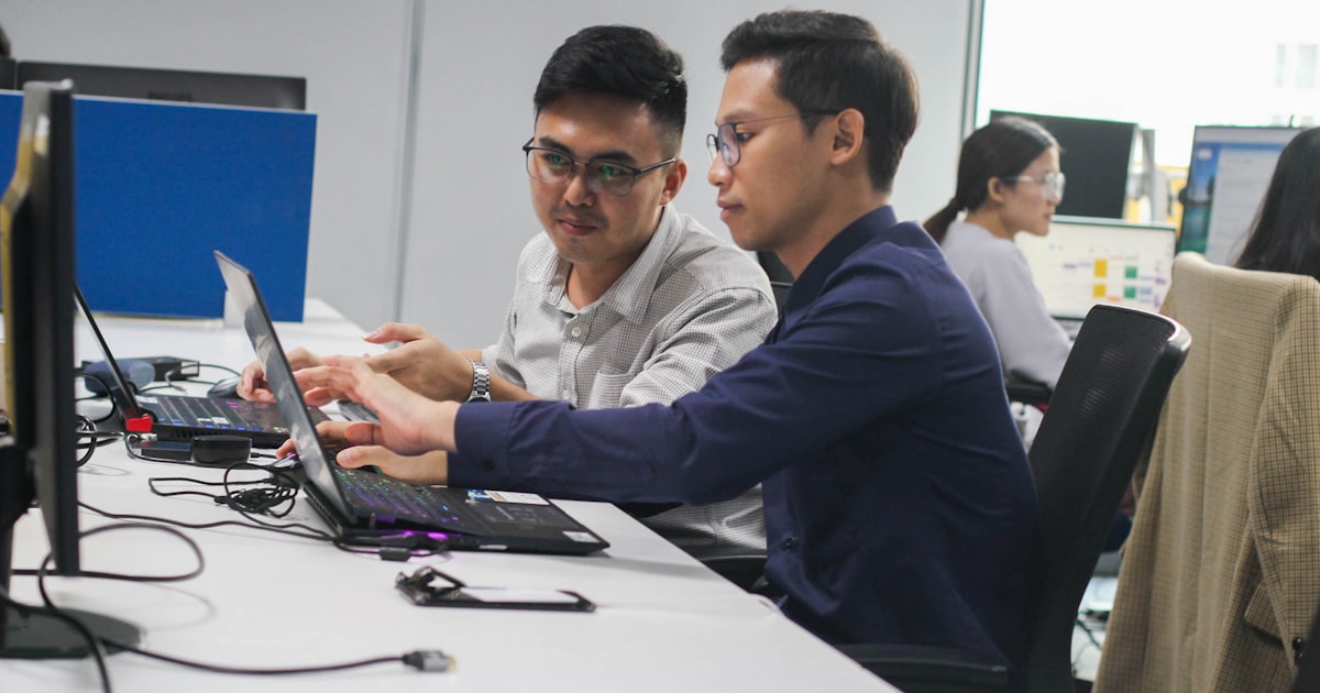 two men sitting at a desk looking at a laptop